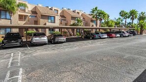 Un parking où plusieurs voitures sont garées devant un immeuble doté de nombreux balcons et bordé de palmiers.