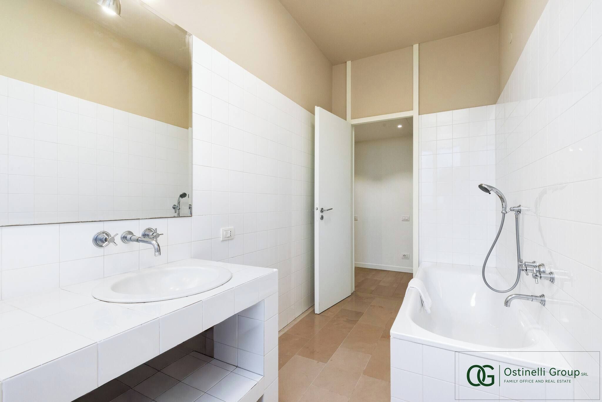 A modern bathroom with two sinks, a large mirror, a bathtub with a showerhead, and a door leading to another room.