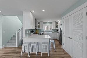 A modern kitchen with white cabinetry, a central island with stools, and a staircase with patterned steps. - Elegant Oceanfront 4 Bedroom 4 Bathroom 14 person home with direct, private beach access and panoramic beach views. (Ocean Isle Beach)