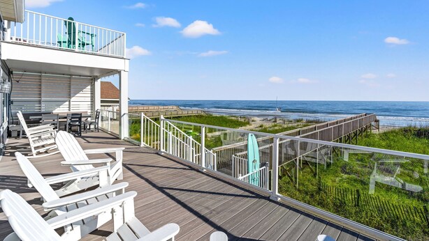 Une terrasse en bois avec des chaises blanches et une table, donnant sur une plage et l’océan.