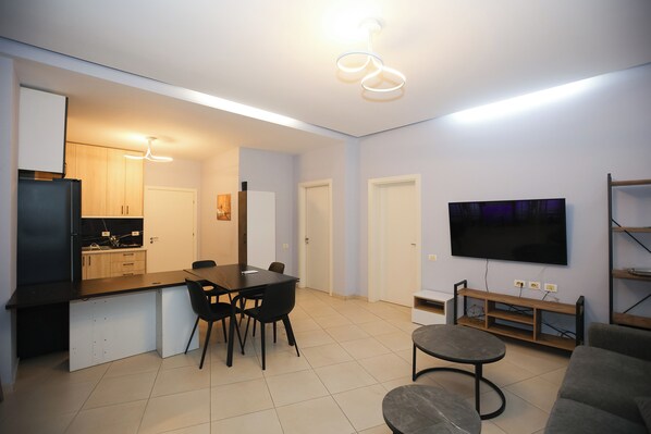 Una sala de estar moderna con un televisor de pantalla plana, un comedor con mesa y sillas, y una cocina con armarios y un refrigerador.