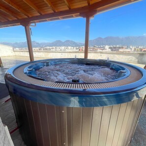 Un bain à remous au revêtement extérieur en bois, entouré d’une pergola en bois, offrant une vue sur un paysage urbain avec des montagnes en arrière-plan.