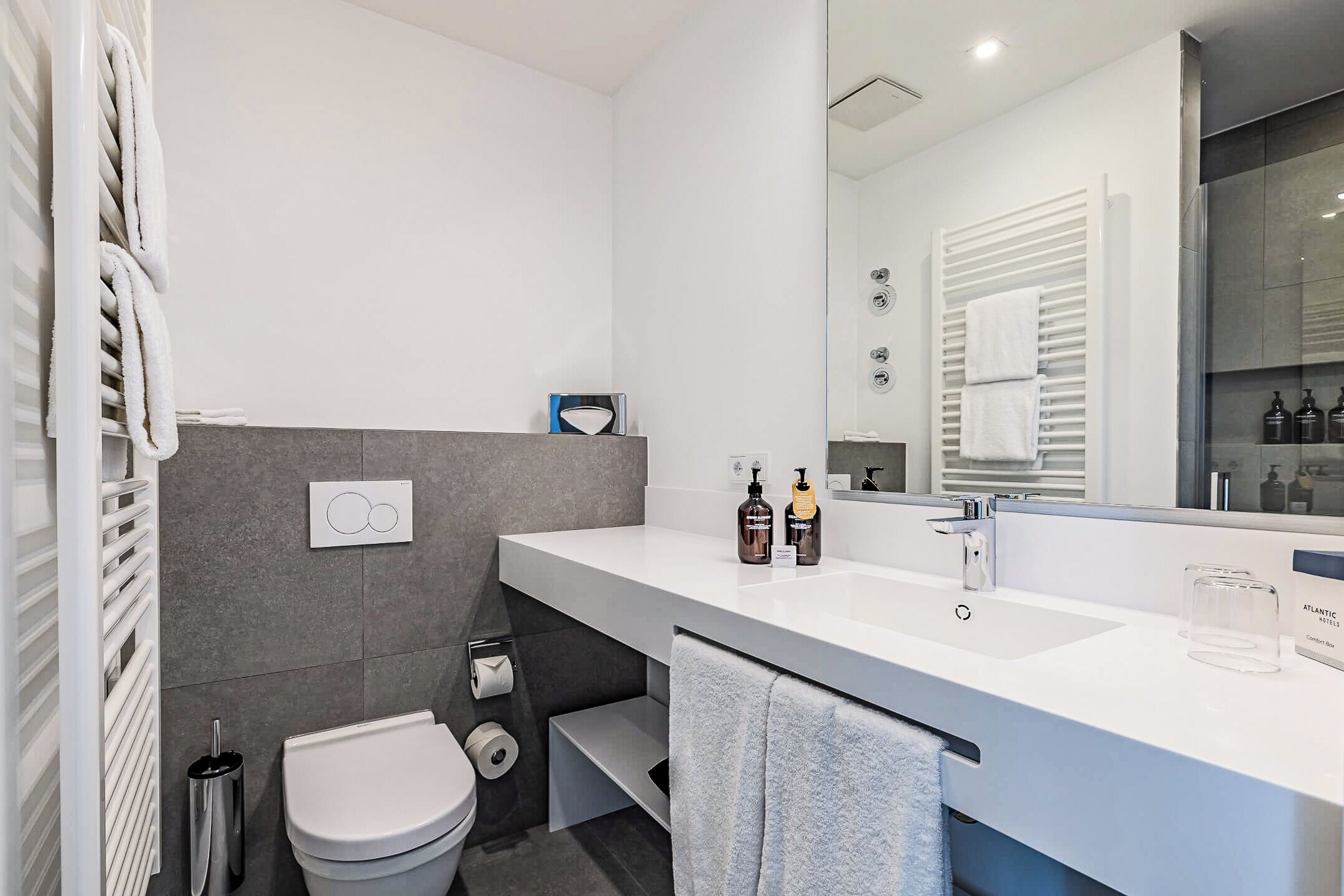 A modern bathroom with a white sink, mirror, and towel rack. There is a toilet with a paper towel holder and a trash can.