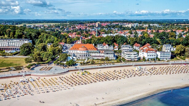 Luchtbeeld van een strand met een pier die in het water reikt, omringd door gebouwen en een heldere lucht.