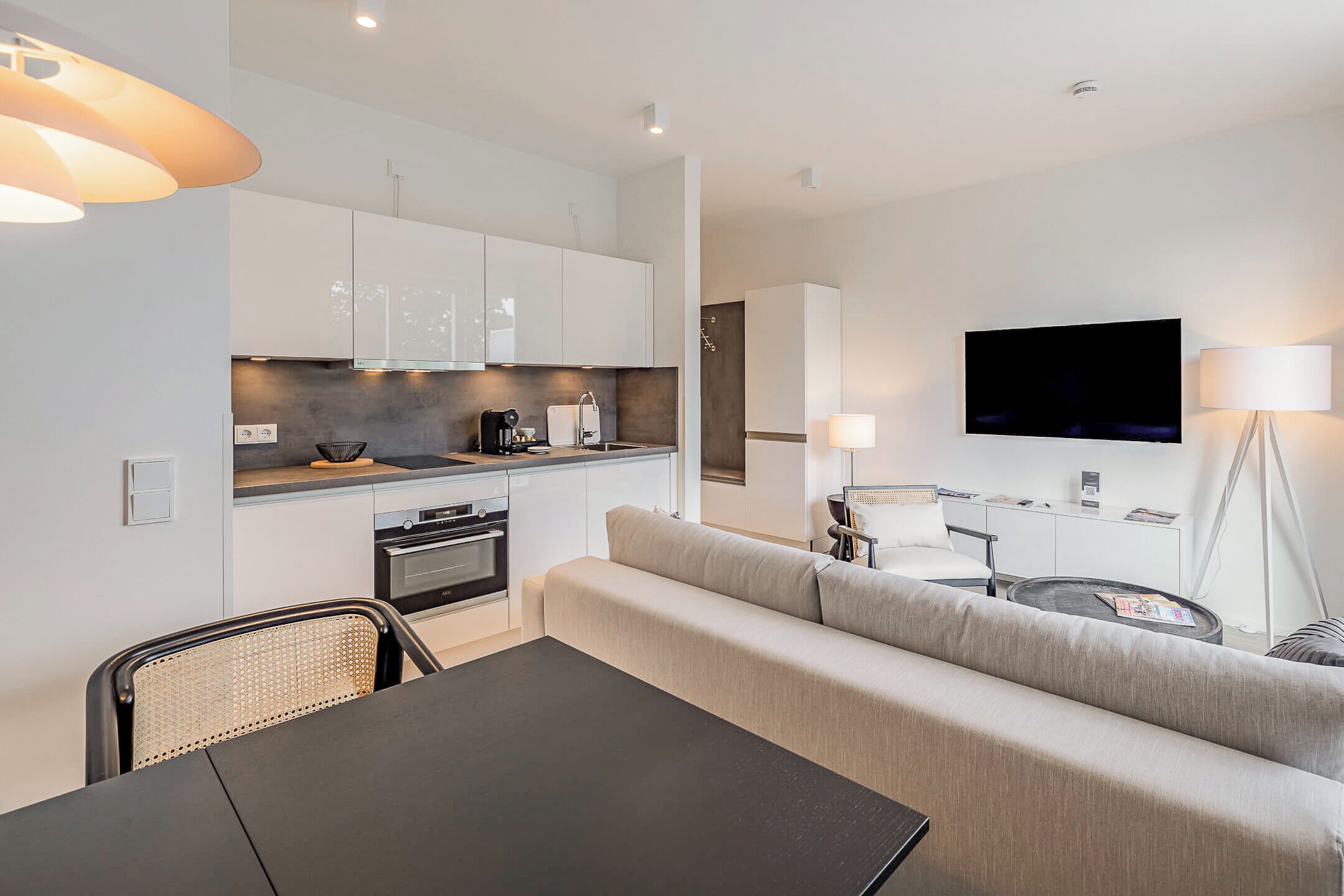 Eine moderne Küche mit weißen Schränken, ein Essbereich mit Tisch und Stühlen, eine Couch und ein an der Wand befestigter Fernseher.