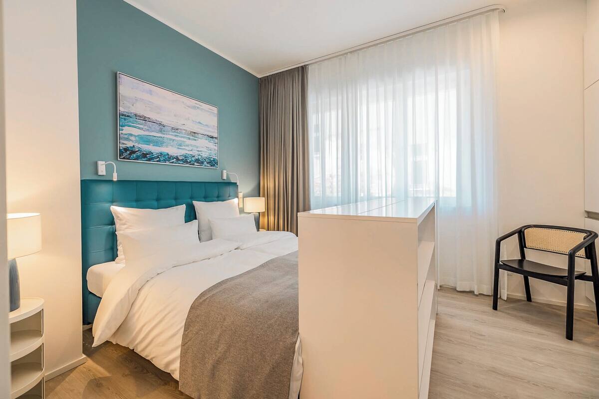 Ein modernes Hotelzimmer mit einer wandfarbenen Akzentwand in Türkis, einem großen Bett mit weißer Bettwäsche, einem Schreibtisch mit Stuhl und einem Fenster mit durchsichtigen Vorhängen.