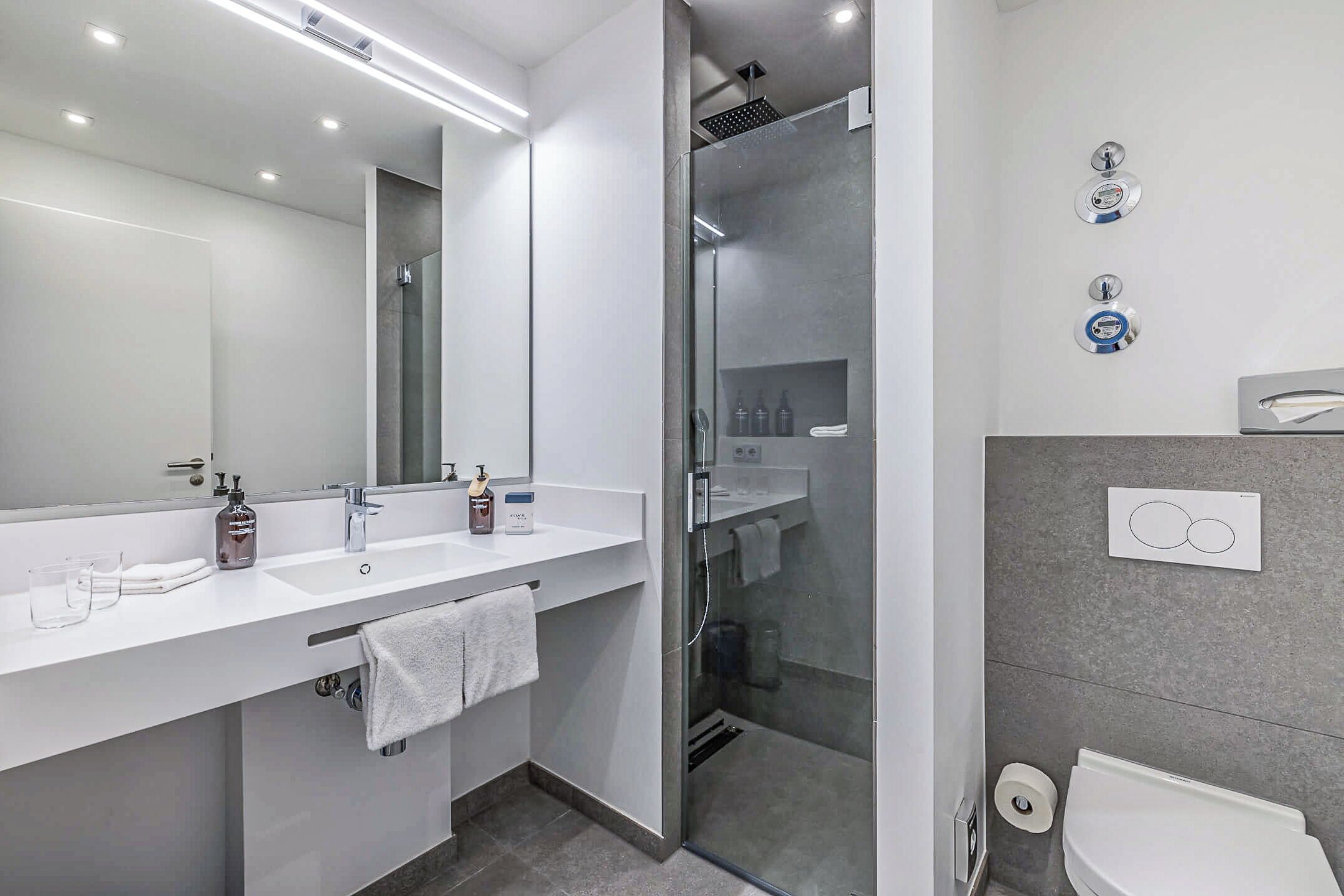 Un baño moderno con un espejo grande, dos lavamanos, cabina de ducha con mampara de cristal y un inodoro.