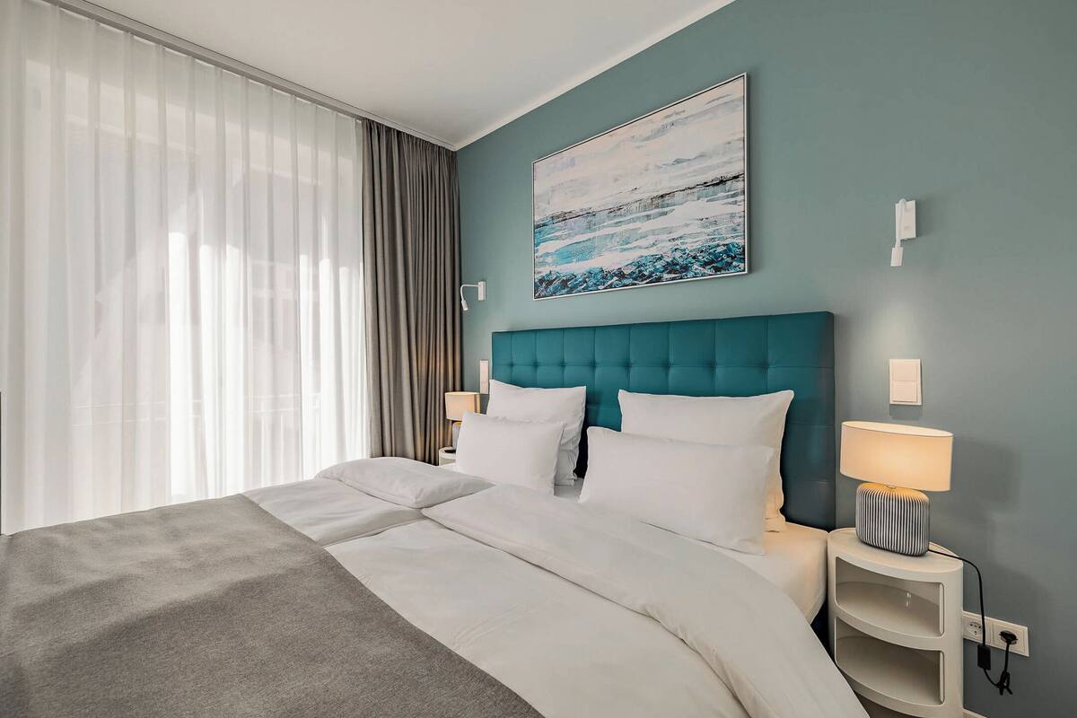 Ein Hotelzimmer mit einem großen Bett, einem blaugrünen Kopfteil, weißer Bettwäsche, einer Nachttischlampe und einem Bild an der Wand.