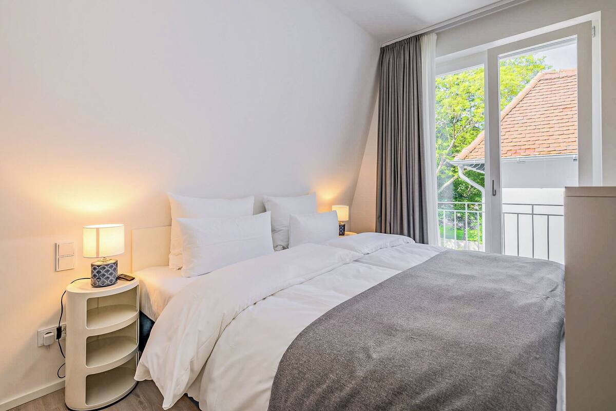 Ein ordentlich bezogenes Bett mit weißer Bettwäsche und einer grauen Decke, ein Nachttisch mit Lampe sowie ein Fenster mit Vorhängen.