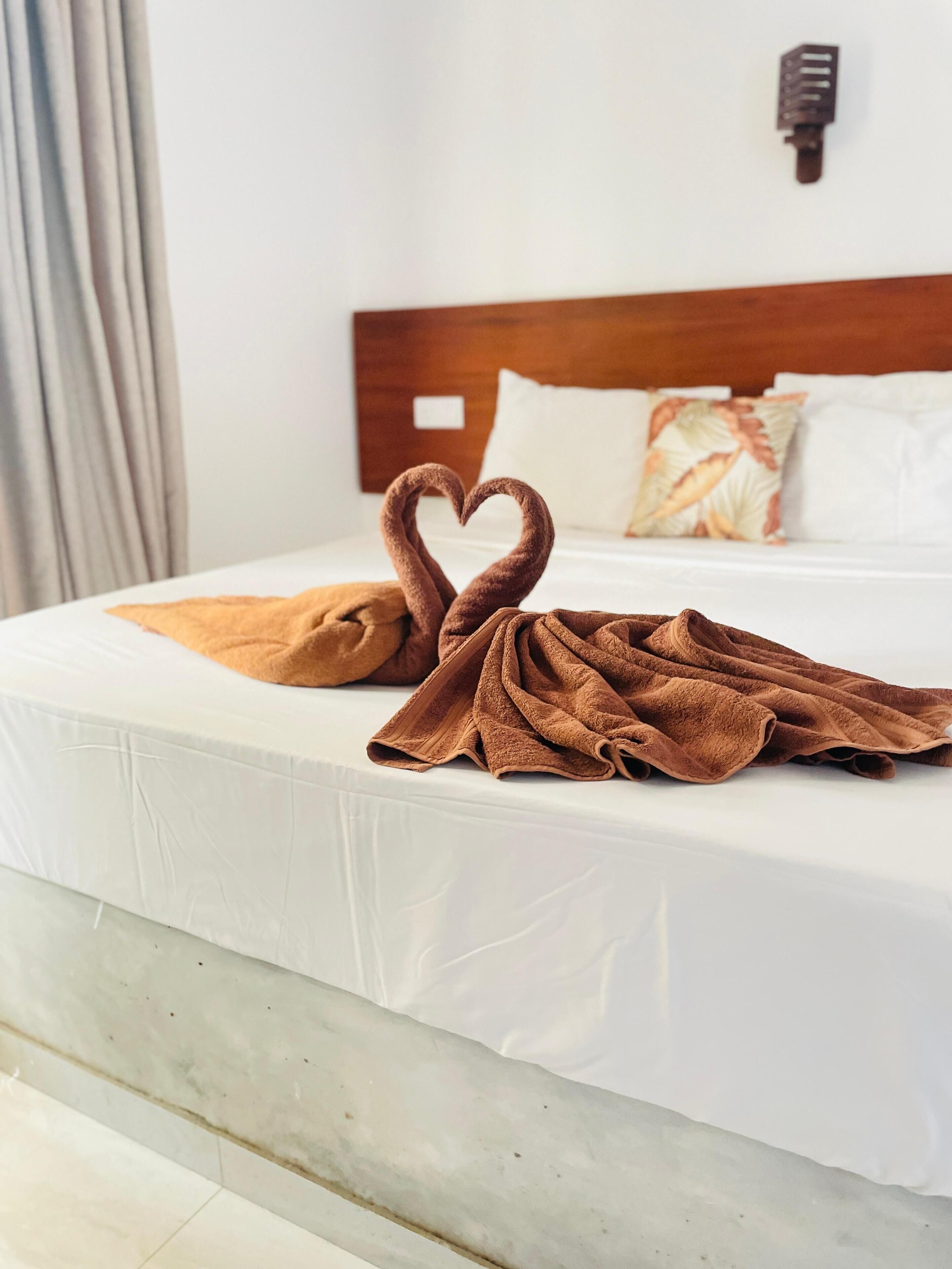 A hotel room with a neatly made bed, a heart-shaped towel arrangement, and a wooden headboard.