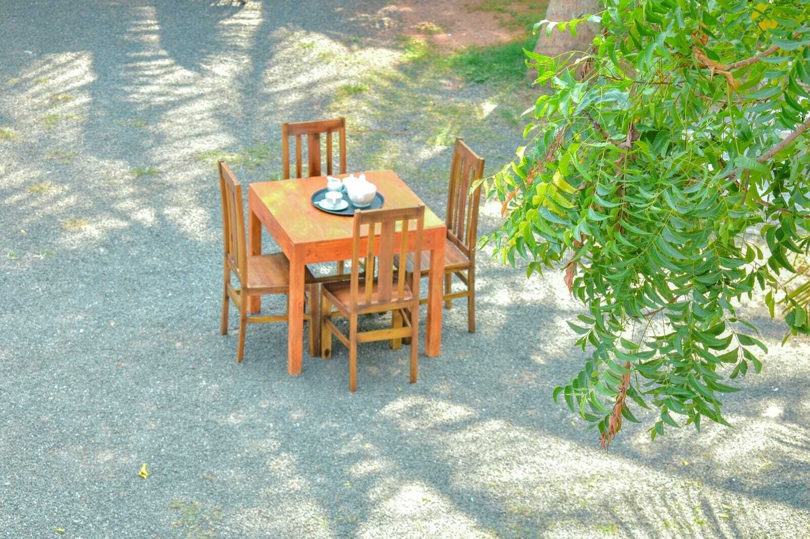 An outdoor wooden table and chairs set on a gravel surface, surrounded by greenery.