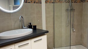 Une salle de bain moderne avec un grand miroir rond, un lavabo, une douche dotée d’une porte en verre et un sol carrelé.