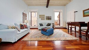 A spacious living room with a white sofa, a blue ottoman, and a patterned rug. There are two doors leading to outdoor areas, and a wooden table with chairs. - Flatinrome Spagna Spanish Steps (ROMA)
