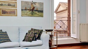 A cozy room with a white sofa, colorful pillows, and a quilt. There are framed paintings on the wall and a view of a building outside through a window. - Flatinrome Spagna Spanish Steps (ROMA)