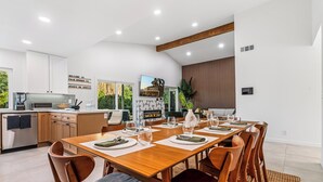 A modern kitchen with white cabinets, a wooden dining table set for six, and a large window overlooking greenery. - Oasis On Arroyo - 3 bed, 2 bath, separate office (Palm Springs)