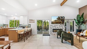 A modern kitchen with a central island, a dining area with wooden chairs, a living room with a fireplace, and a large potted plant. - Oasis On Arroyo - 3 bed, 2 bath, separate office (Palm Springs)