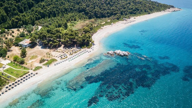 Una vista aerea di una zona costiera con spiaggia, acque blu cristalline e una collina ricoperta di boschi.