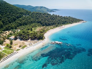 An aerial view of a coastal area with a beach, clear blue waters, and a forested hillside.