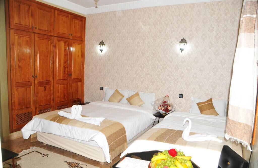 Une chambre d’hôtel avec deux lits, une armoire en bois, des appliques murales et un panier de fruits posé sur le lit.