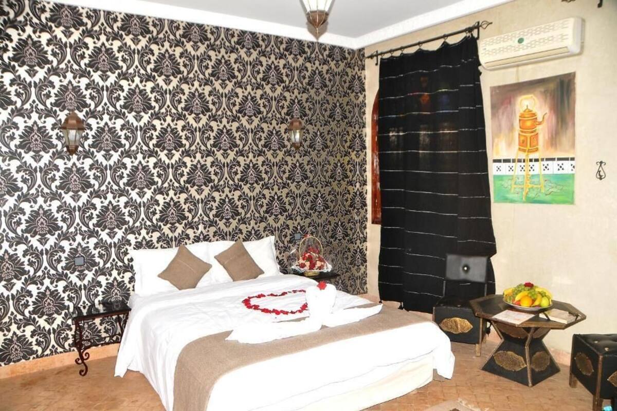 A hotel room with a patterned wallpaper, a bed with white linens, a nightstand with a lamp, a black curtain, and a wall-mounted air conditioner.
