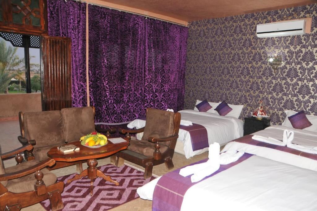 Una camera d'albergo con due letti, una sedia di legno, un tavolino con frutta e tende viola.