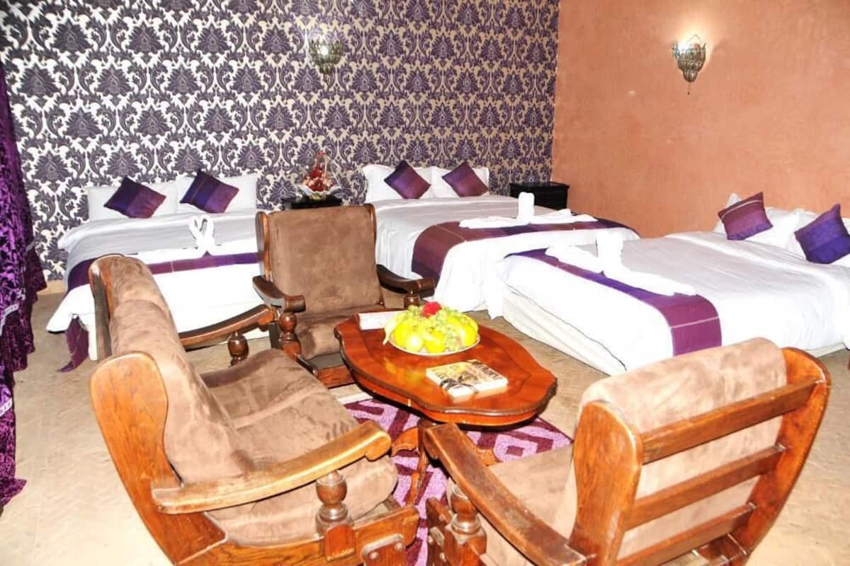 A hotel room with two single beds, a sofa, a wooden coffee table with fruit, and patterned wallpaper.