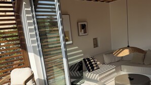 Een moderne buitenruimte met een houten pergola, een glazen deur naar een balkon, een bank, een rond poefje en een geweven kussen.