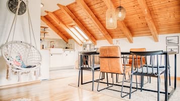 Una habitación estilo loft con vigas de madera a la vista, un lámpara colgante, un sillón colgante de macramé y una zona de comedor con una mesa y sillas.