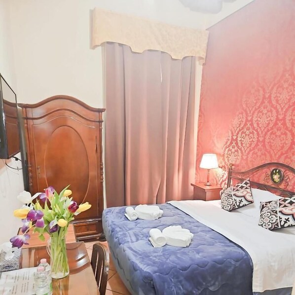 Una camera d'albergo con un poggiatesta in legno, un letto con copriletto blu, un comodino con una lampada e un vaso di fiori sulla scrivania.
