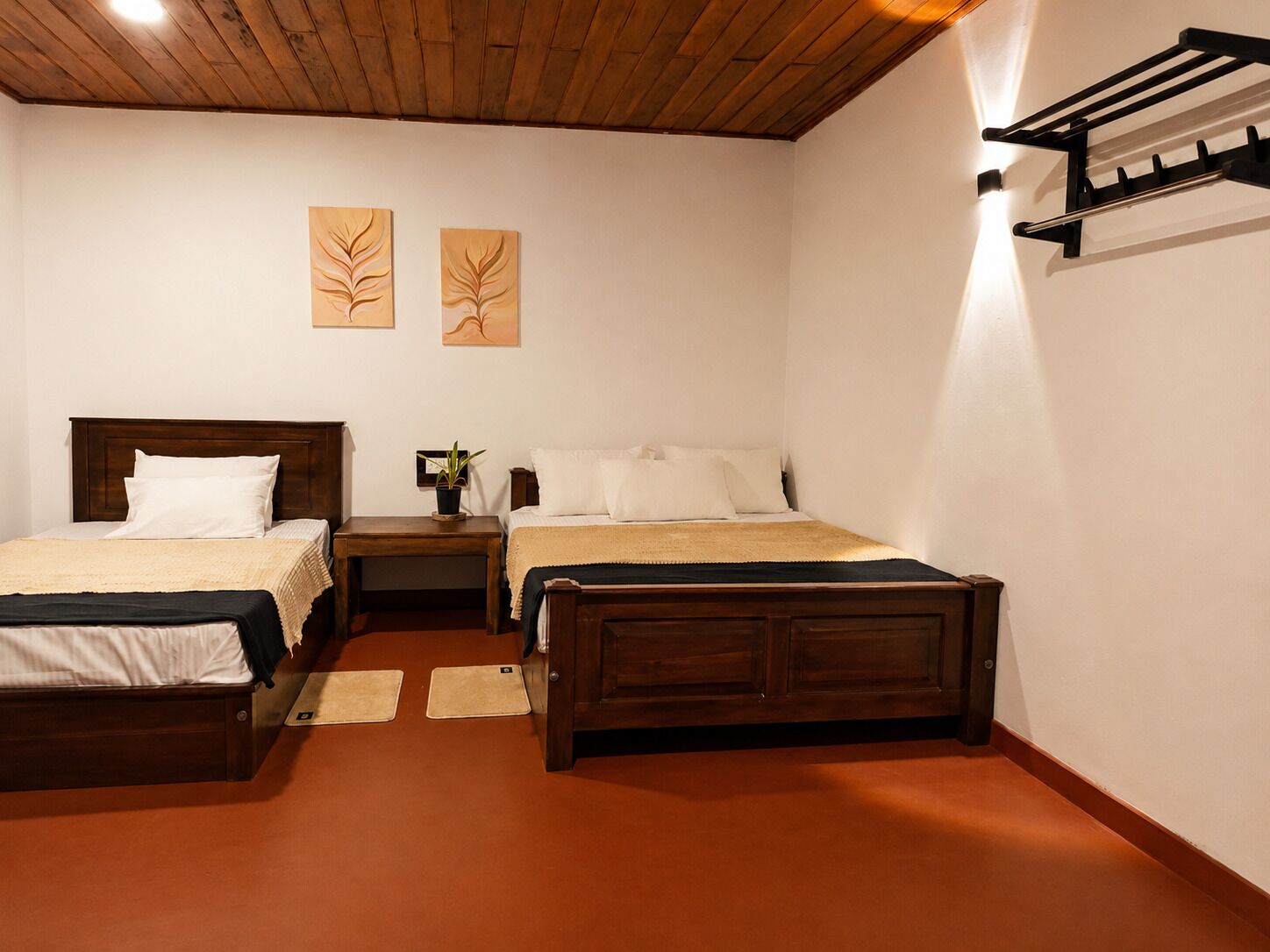 A room with two beds, a wooden headboard, a nightstand with a plant, and a wall-mounted rack.