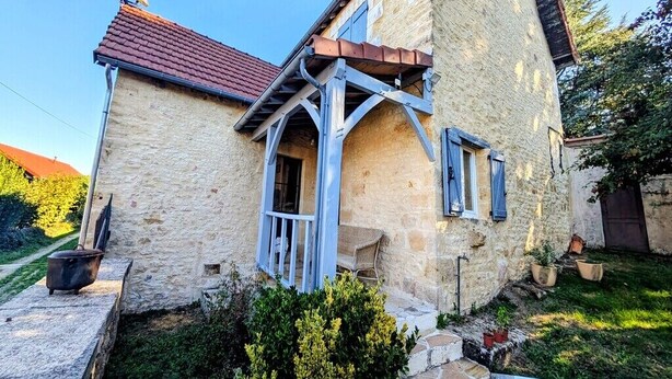 A stone building with a red-tiled roof, a wooden porch, and a small garden area with potted plants.