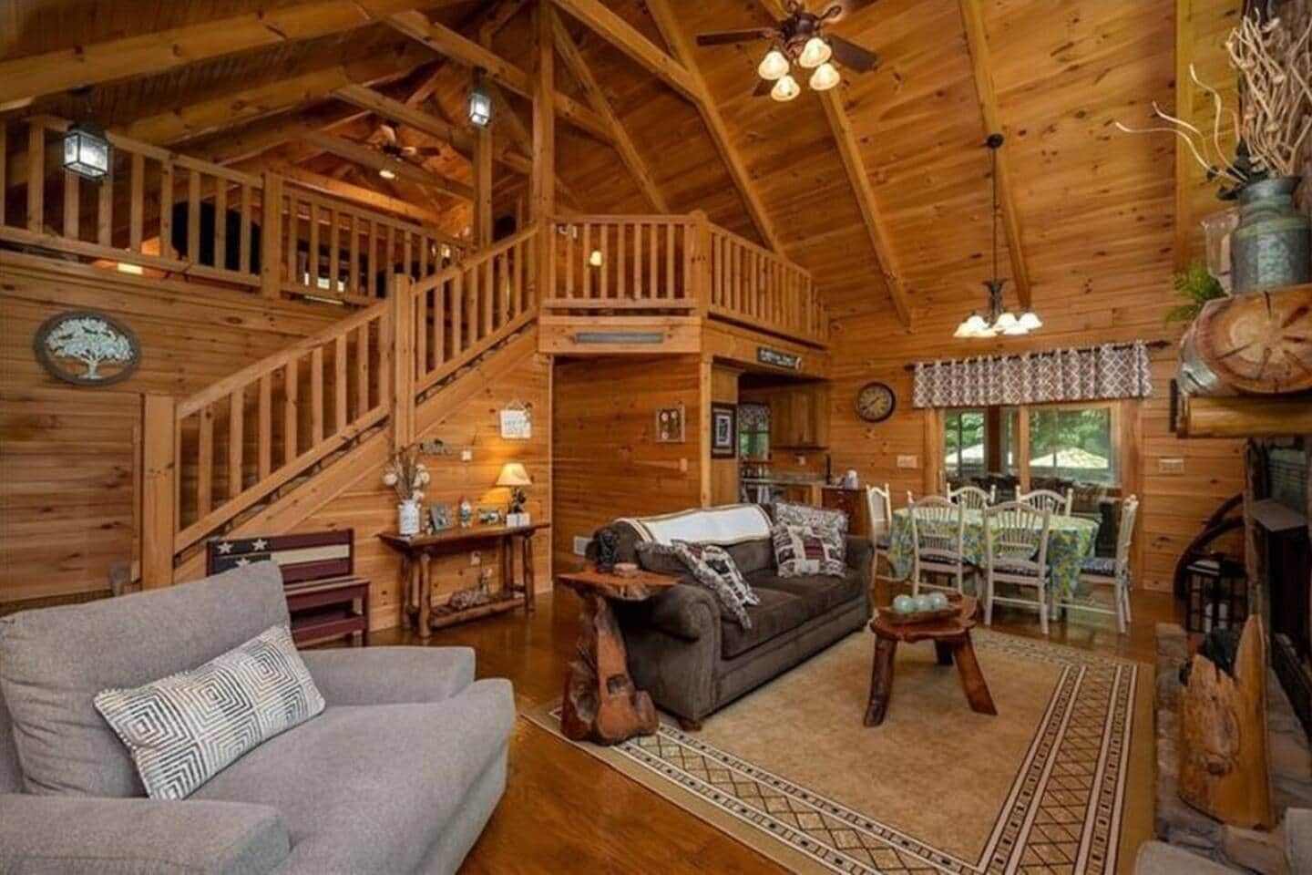 A cozy cabin interior with a wooden staircase, a fireplace, a dining area, and a living room furnished with a sofa and a coffee table.