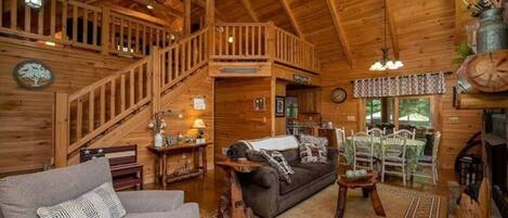A cozy cabin interior with a wooden staircase, a fireplace, a dining area, and a living room furnished with a sofa and a coffee table.