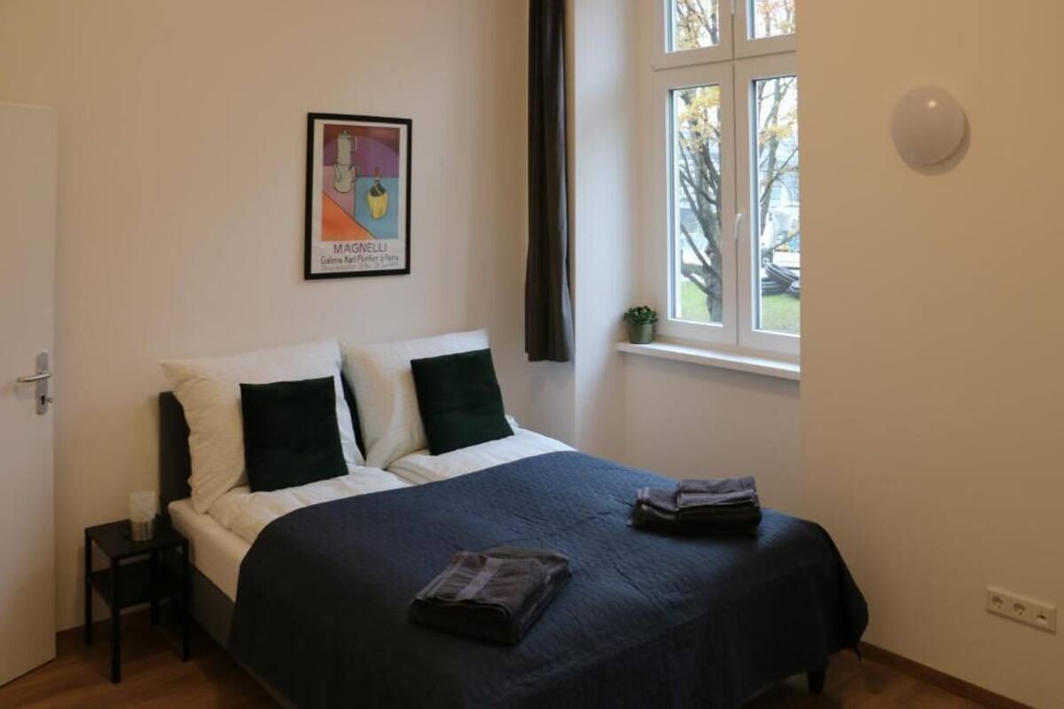 A neatly made bed with a dark blue bedspread and two pillows, a small bedside table with a lamp, a framed artwork on the wall, and a window with a view of trees and a car outside.