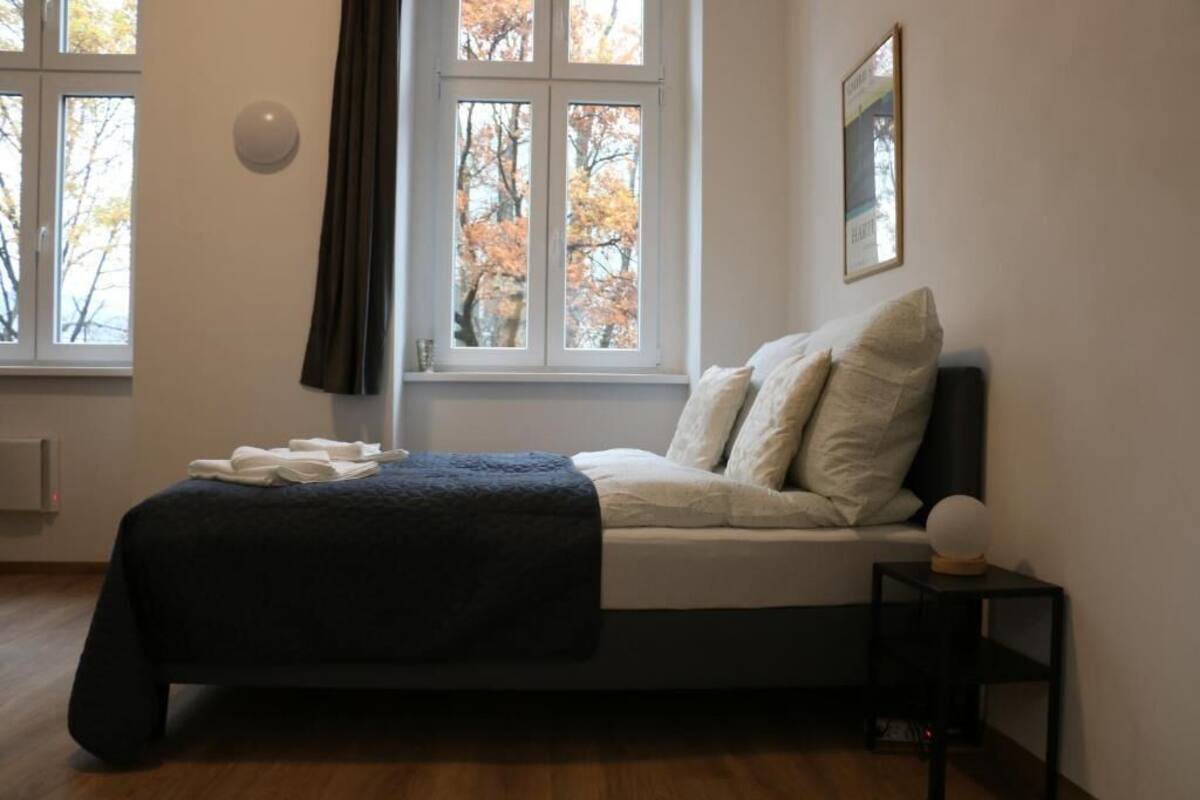 A bedroom with a bed, a window with curtains, a bedside table with a lamp, and a framed picture on the wall.