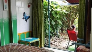 Une pièce comprenant un lit, une table et une chaise. On y trouve une porte-fenêtre coulissante qui donne sur un espace extérieur agrémenté de plantes et d’une chaise rouge.