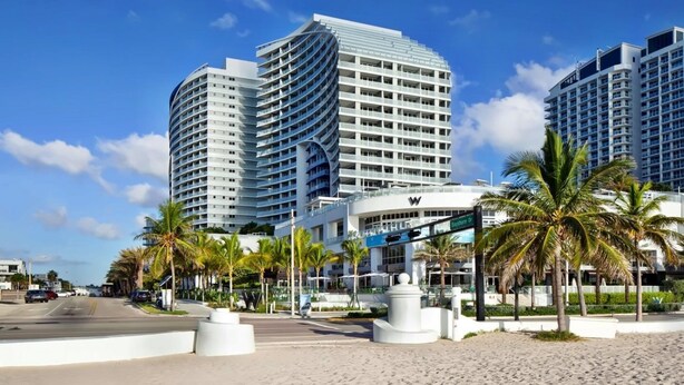Un hôtel moderne à la façade incurvée, situé en bord de mer, avec des palmiers et un ciel dégagé.