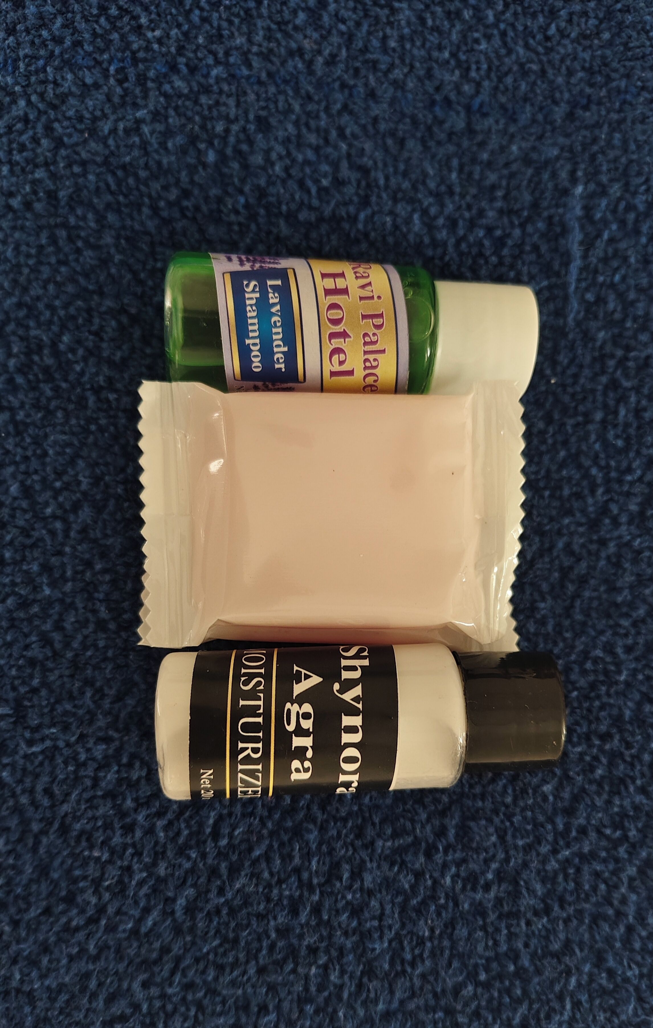 Three toiletry items: a green bottle labeled 'Lavender Shampoo,' a beige bottle labeled 'Hotel Palace,' and a black bottle labeled 'Shynor Agra.'