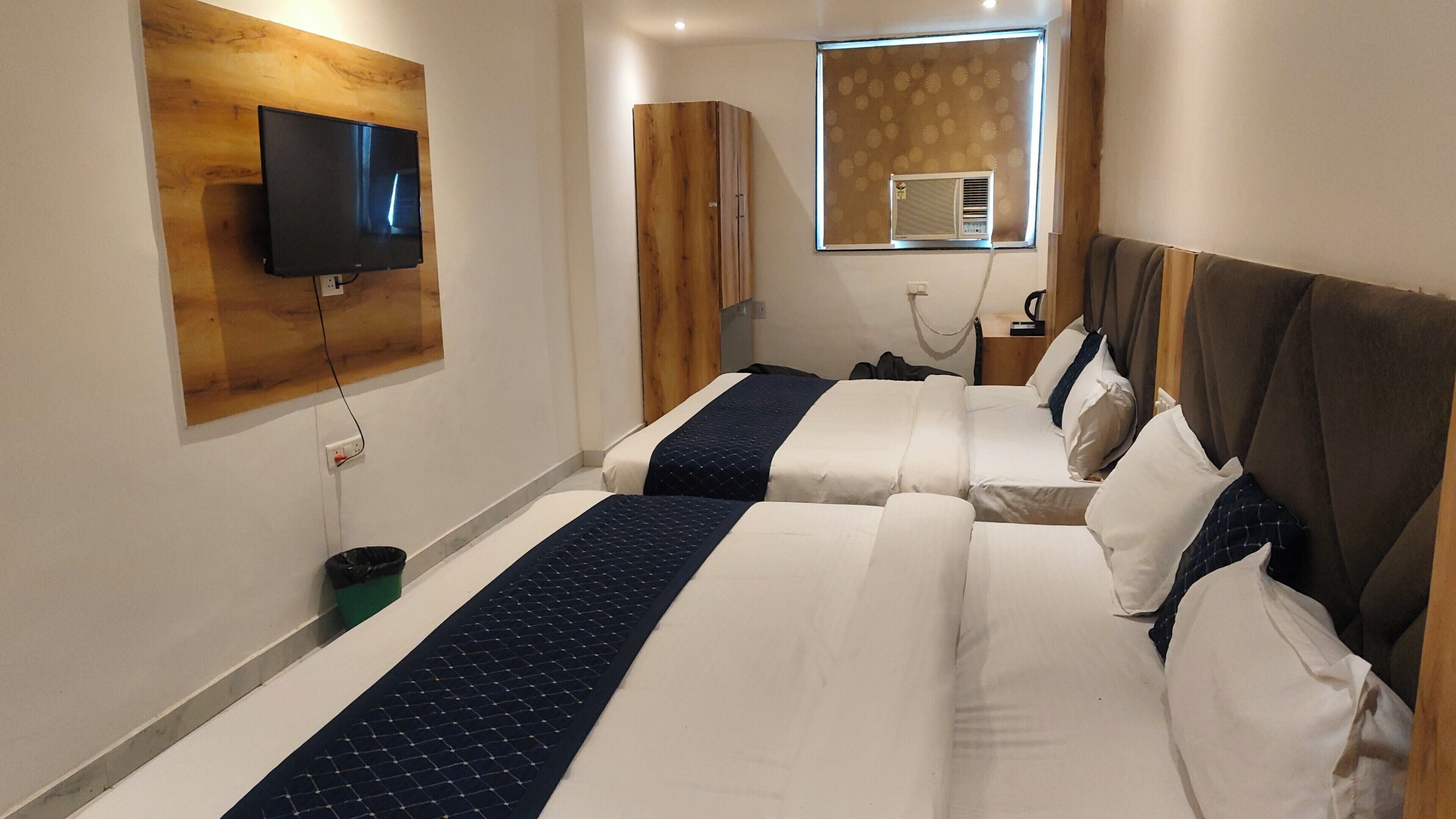 A hotel room with two beds, a TV mounted on a wooden panel, a window with an air conditioner, and a nightstand with a lamp.