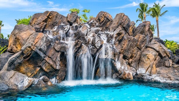 Ein Wasserfall stürzt über große Felsen in ein klares, blaues Becken, umgeben von Palmen.