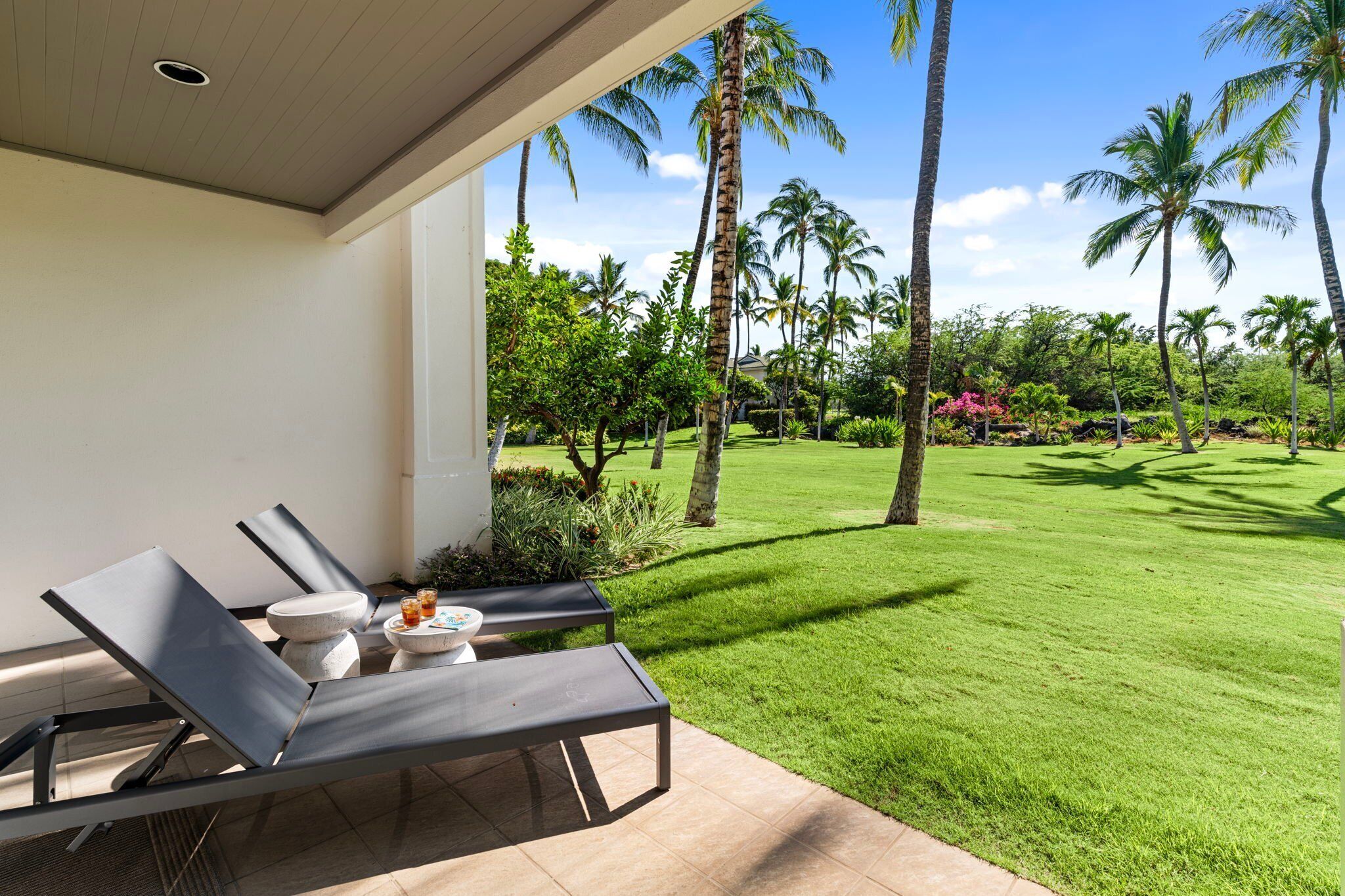 A patio with two lounge chairs and a small table, overlooking a well-manicured lawn with palm trees and a clear blue sky.