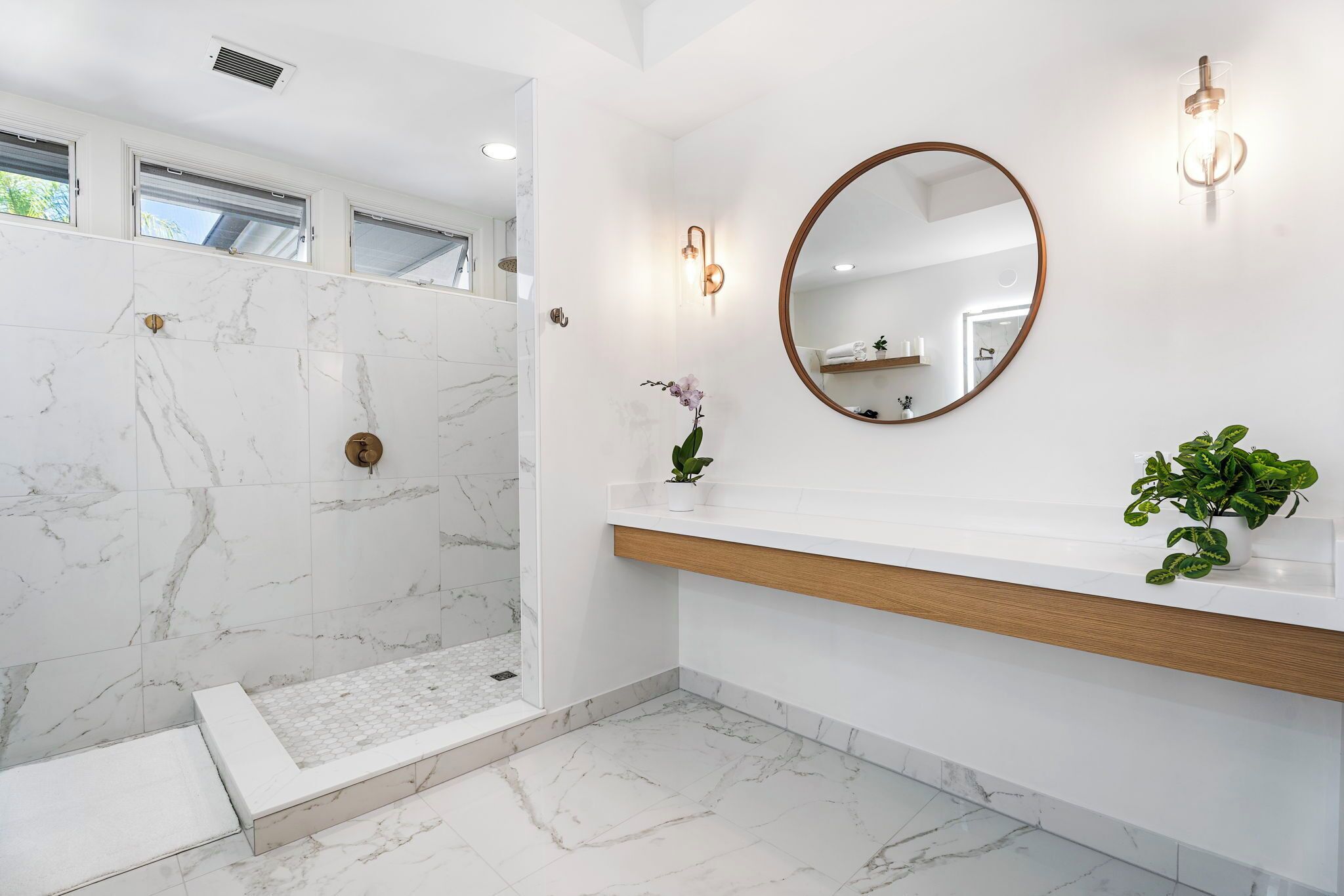 A modern bathroom with a walk-in shower, marble flooring, a wooden vanity, a round mirror, and wall-mounted sconces.