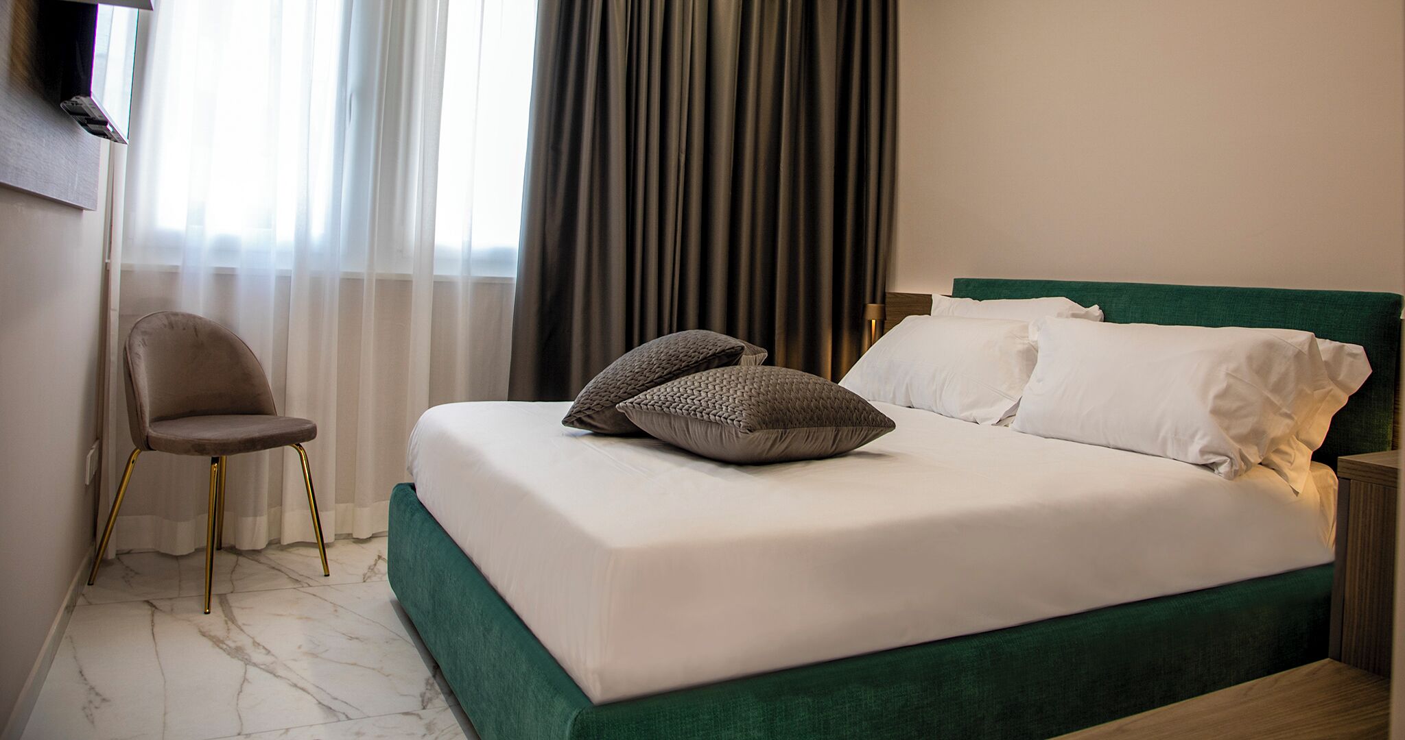 Un lit bien fait, avec des oreillers blancs et gris, une tête de lit recouverte d’un tissu vert et un sol en marbre.