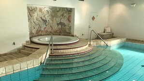 En pool med en rund bubbelpool, en stenmur med abstrakt konst och en liten sittgrupp med en kudde.