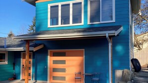 Una casa de dos pisos con fachada azul, una puerta principal, un garaje y un pequeño porche con dos sillas.