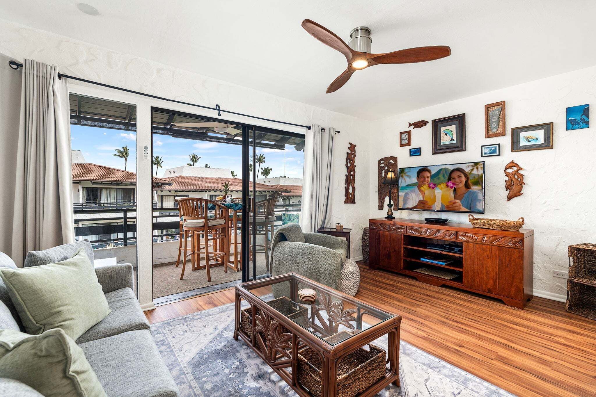 A living room with a sofa, a glass coffee table, a wooden entertainment unit, and a balcony with a view of palm trees.