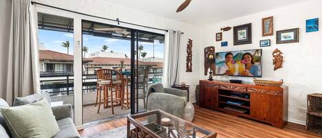 Un salon comprenant un canapé, une table basse en verre, un meuble de rangement en bois et un balcon offrant une vue sur les palmiers.