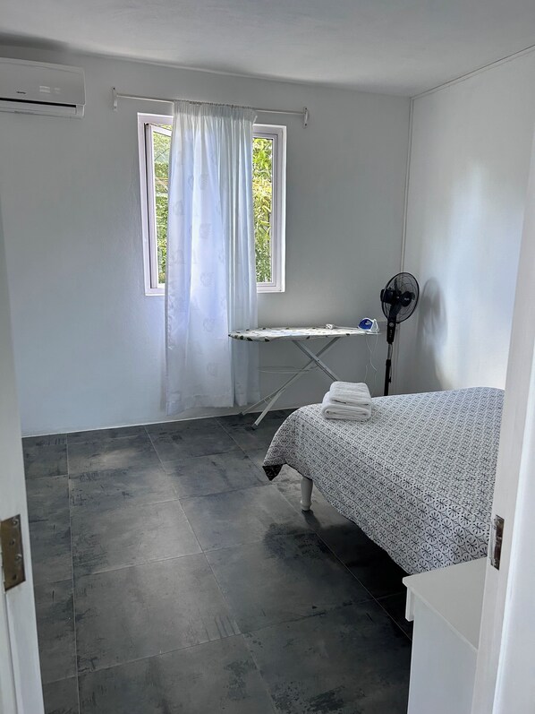A room with a bed, an ironing board, a fan, and a window with curtains.