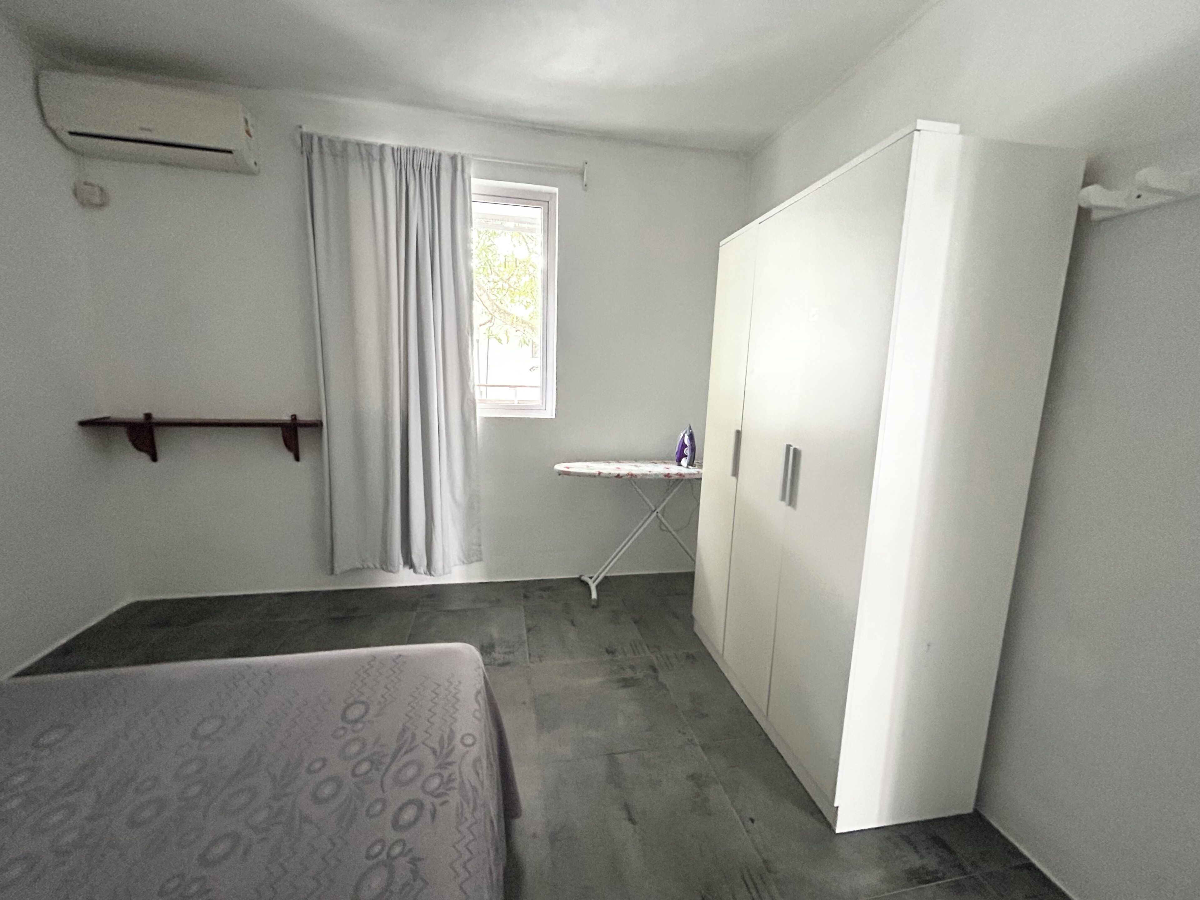 Ein Zimmer mit einem Bett, einem Schrank, einem kleinen Tisch mit Bügeleisen und einem Fenster mit Vorhängen.