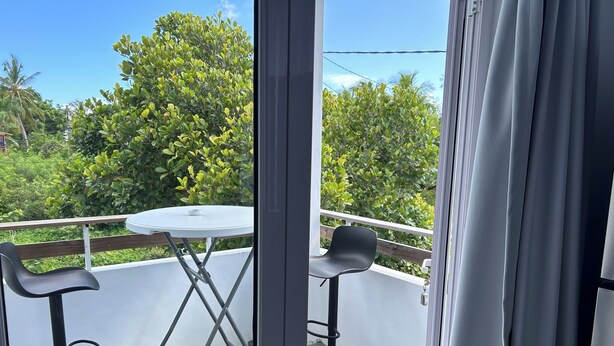 Ein Balkon mit einem runden Tisch und zwei schwarzen Stühlen, einem weißen Geländer sowie Blick auf grüne Bäume und einen klaren Himmel.
